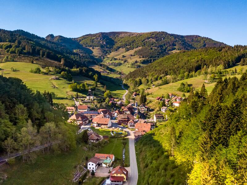 Eine malerische Aussicht auf ein kleines Dorf inmitten einer grünen Hügellandschaft mit dichten Wäldern und einem klaren blauen Himmel im Hintergrund.
