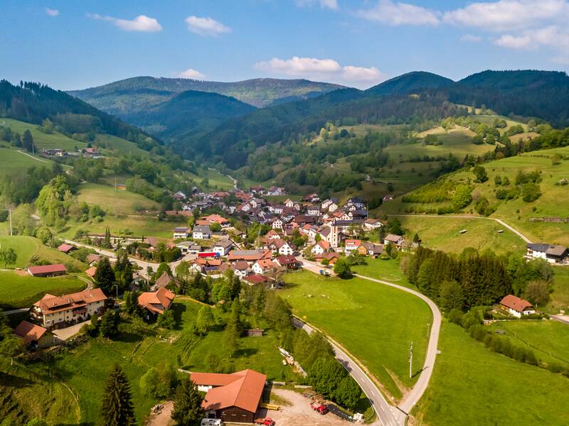 Eine idyllische Dorfansicht mit verstreuten Häusern und grünem, von bewaldeten Hügeln umgebenem Hügelland unter blauem Himmel.