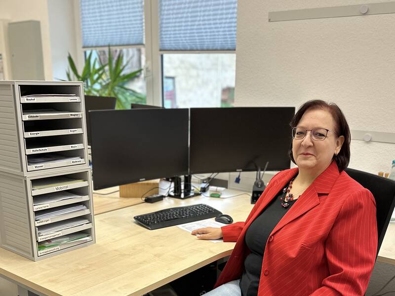 Eine Frau in rotem Blazer sitzt an einem Büroarbeitsplatz mit zwei Monitoren und einem Stapel Ablagefächer.